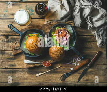 Des hamburgers avec bacon croustillant et légumes frais dans de petites casseroles en fonte et verre de bière de froment sur conseil rustique ove Banque D'Images