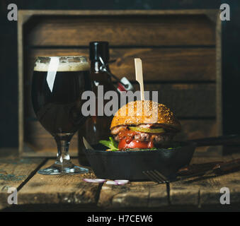 Burger de boeuf maison avec bacon croustillant, des légumes frais et du ketchup dans poêle en fonte noire servi avec un verre de bière brune sur ru Banque D'Images