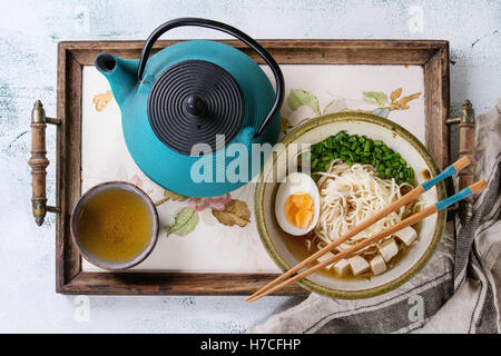 Bol de soupe avec des nouilles de style asiatique, l'oeuf, l'oignon de printemps et le tofu, fromage servi avec des baguettes, théière et tasse de thé vert sur Banque D'Images