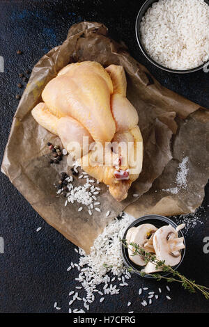 Mini cru entier poulet sur du papier sulfurisé avec assaisonnement, le thym, les champignons tranchés et bol de riz blanc non cuit sur fond noir textur Banque D'Images