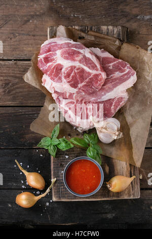 Steak de porc cru viande cou sur planche à hacher l'oignon, l'échalote avec la sauce tomate, le basilic et l'assaisonnement de la marinade, servi avec fourchette à viande Banque D'Images