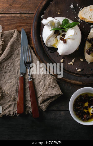 Tranches de fromage italien burrata servie sur planche à découper ronde avec les pignons de pin, des tranches de pain, basilic et tomates séchées dans l'huile d'olive Banque D'Images