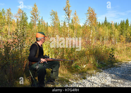 Chasseur d'orignal à capuchon orange assis sur une chaise tenant un fusil, photo du nord de la Suède. Banque D'Images