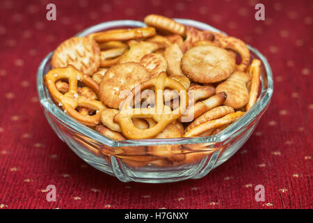 Mélange de snacks salés (crackers et bretzels) dans bol en verre sur fond rouge Banque D'Images