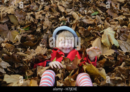 Professionnels de l'automne. Petite fille en manteau rouge jouant avec pile de feuilles sèches à l'automne parc. Banque D'Images