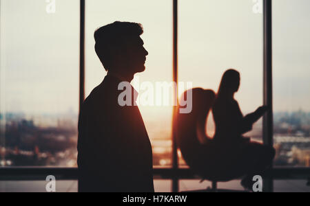 Silhouette de jeune homme barbu élégant en costume officiel, debout devant la fenêtre et son collègue Banque D'Images