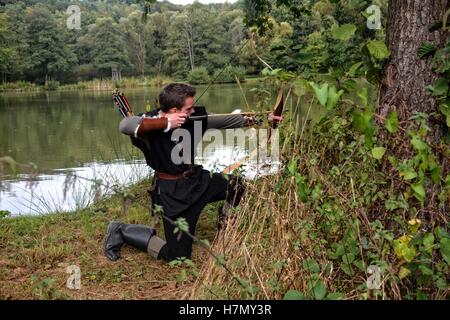 Archer médiéval avec capot noir s'agenouille sur le côté sur le terrain avant un lac, vise avec la flèche et la courbe de côté Banque D'Images