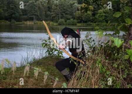 Archer médiéval avec capot noir s'agenouille sur le sol avant d'un lac, vise avec la flèche et la courbe vers l'avant Banque D'Images