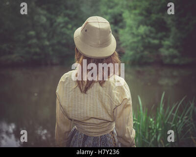 Une jeune femme portant un chapeau de safari est debout par un étang dans la forêt sur une journée ensoleillée Banque D'Images