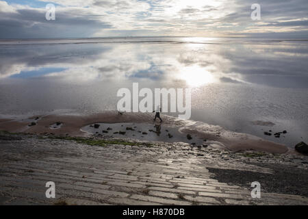 Une femme et son chien à pied le long de la plage avec le ciel reflété sur la surface humide de l'estuaire à marée basse (hiver) Banque D'Images