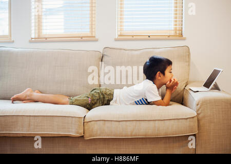 Maison de famille. Un garçon couché sur un canapé devant une tablette numérique. Banque D'Images