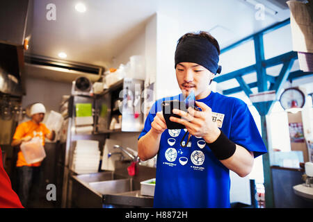 La boutique de nouilles ramen, le personnel de préparer des aliments. Un chef en utilisant un téléphone intelligent dans une cuisine. Banque D'Images