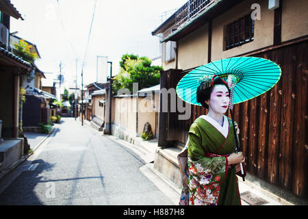 Une femme vêtue de la traditionnelle style geisha en kimono, tenant un parasol en papier marchant le long d'une rue. Banque D'Images