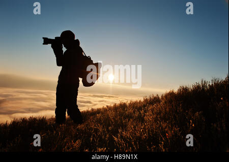 Silhouette of man photographe avec appareil photo sur les montagnes en arrière-plan la main sur le coucher du soleil avec brouillard. Amazing shot of beauty world et hum Banque D'Images