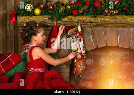 La princesse d'hiver - cute little girl in red dress mettre des cadeaux dans les chaussettes, suspendu à une cheminée. Se félicite de la nouvelle année, noël Banque D'Images