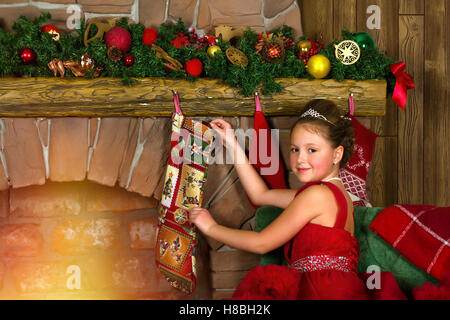 La princesse d'hiver - cute little girl in red dress mettre des cadeaux dans les chaussettes, suspendu à une cheminée. Se félicite de la nouvelle année, noël Banque D'Images