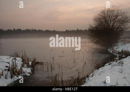 Lac d'hiver couvert de glace au coucher du soleil alors que le brouillard se lève sur le lac, une forêt en arrière-plan Banque D'Images