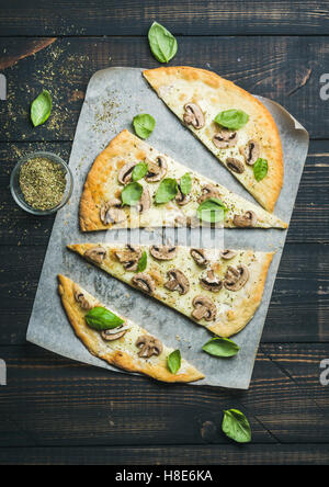 Pizza aux champignons fait maison avec du basilic coupé en tranches et les épices en verre sur du papier sulfurisé sur fond de bois brûlée sombre, top v Banque D'Images