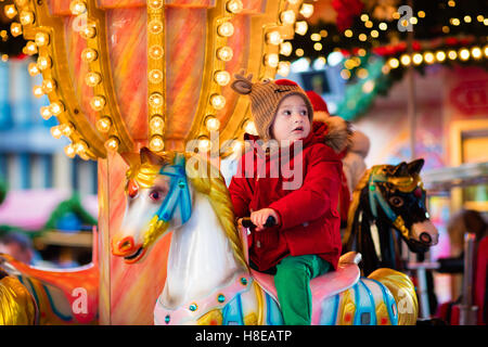 Heureux petit garçon en veste chaude et rouge nordique tricoté bonnet et écharpe riding carousel horse au cours de voyage en famille Banque D'Images