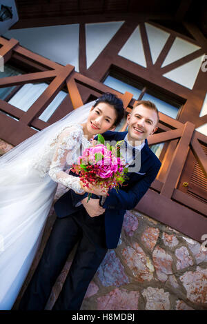 Low angle portrait of happy wedding couple holding flower bouquet contre house Banque D'Images