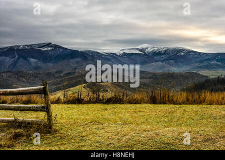Clôture en bois sur le pré vert, mais avec de l'herbe congelé en face de la plage de haute montagne aux cimes enneigées sur un jour d'automne nuageux Banque D'Images