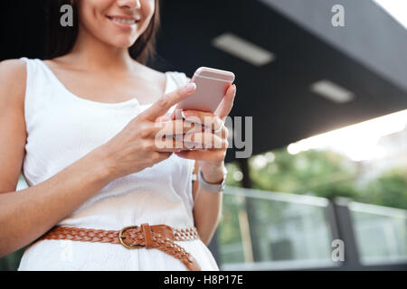 Smiling jolie jeune femme debout et en utilisant smartphone outdoors Banque D'Images
