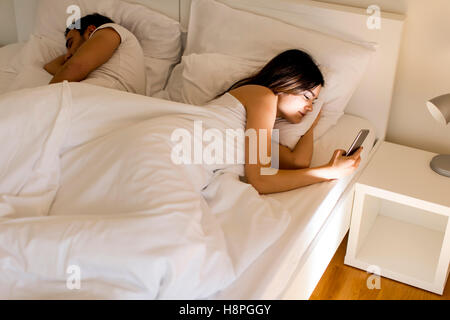 Jeune couple au lit. L'homme dort tandis qu'une jeune femme à la nuit au téléphone Banque D'Images