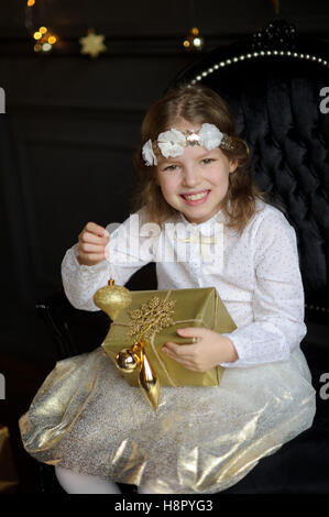 Cheerful girl de 8-9 ans est assis dans une chaise avec de magnifiques paniers fort dans les mains. La fille est habillée d'une robe or blanc avec un wreat Banque D'Images