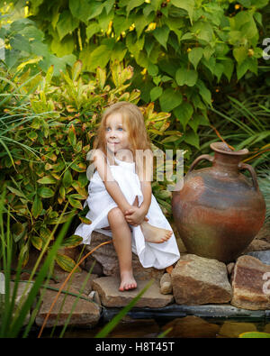 Jolie petite fille en robe blanche assise à l'étang avec des nénuphars. Ancien pot en argile debout à côté d'elle. Banque D'Images