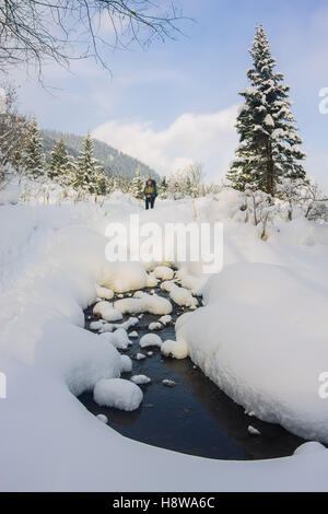 Backpacker debout devant un ruisseau qui coule à travers l'hiver les dérives de la neige et des arbres. Banque D'Images