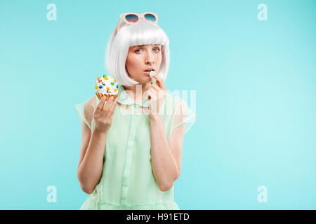 Belle jeune femme en perruque blonde et manger debout sur fond bleu cupcake Banque D'Images