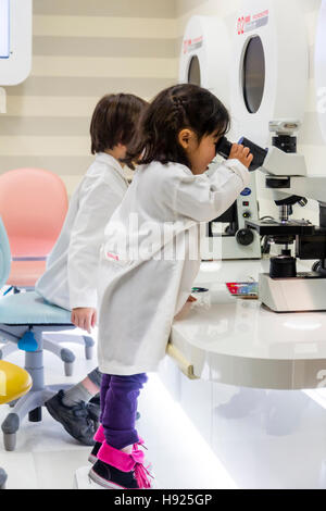 Le Japon, Nishinomiya, KidZania. Les enfants, garçon et fille en blanc blouses de laboratoire, à l'aide de microscopes, petite fille sur la pointe des pieds l'effort pour voir. Banque D'Images