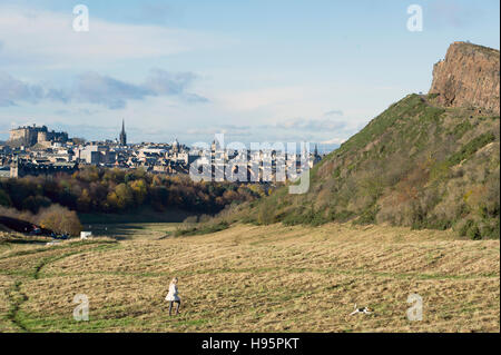 Une jeune femme marche son chien à Holyrood Park avec le radical road sur Salisbury Crags dans la distance. Banque D'Images