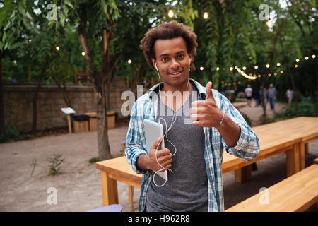 Heureux jeune homme africain des écouteurs holding tablet et showing Thumbs up Banque D'Images