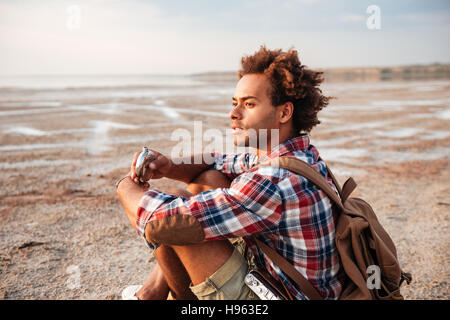 Jeune homme africain pensive avec sac à dos assis et boire du hip flask Banque D'Images