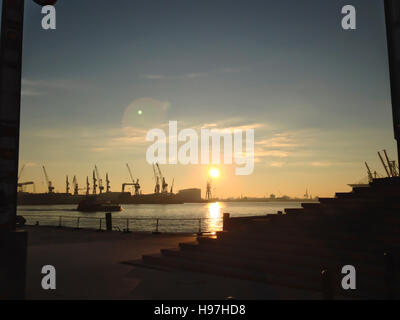 Coucher de soleil sur un port et les quais avec le soleil qui reflète sur l'eau et des grues industrielles silhouetté contre le ciel coloré Banque D'Images