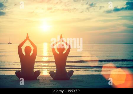Deux silhouettes man and woman practicing yoga au coucher du soleil sur la plage. Banque D'Images