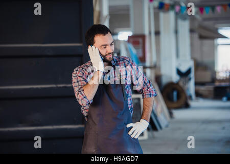 Homme barbu souriant portant des gants, propriétaire d'une petite entreprise de menuiserie, debout dans son atelier studio, talking on mobile phone Banque D'Images