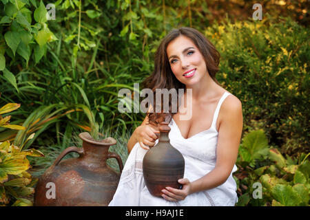 Belle jeune fille en robe blanche tenant une petite vieille cruche. Grand vieux pot en argile debout à côté d'elle. Banque D'Images