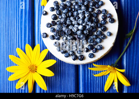 Blueberry cheesecake crémeux sucré avec de petits fruits bleu sur un fond en bois bleu avec des fleurs jaunes. vue d'en haut. Banque D'Images