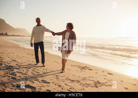 Longueur totale de plein air enjoué senior couple marchant le long de la plage au coucher du soleil. Homme mature et femme marche sur le bord de la mer. Banque D'Images