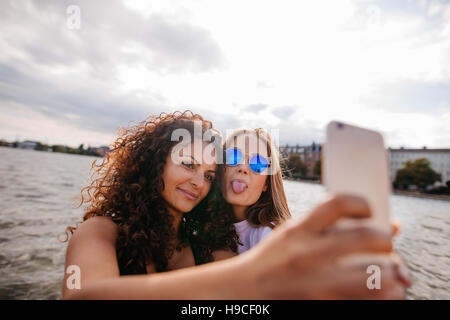 Tourné d'adolescentes en tenant avec selfies smart phone par le lac. Un sticking out tongue avec d'autres holding mobile phone. Banque D'Images