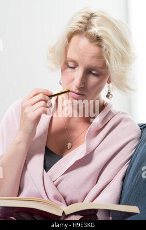 Une femme blonde d'âge moyen habillé est assis sur un canapé ou un canapé bleu tenant un livre et de mordre l'extrémité d'un crayon dans la concentration qu'elle la lit. Banque D'Images
