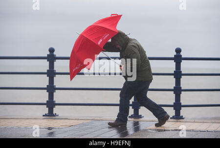 Un homme avec des vents violents combats un parapluie rouge pendant les Angus dans Porthcawl, dans le sud du Pays de Galles, Royaume-Uni. Banque D'Images