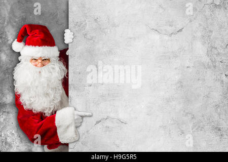 Rouge Blanc classic traditionnel du père Noël en regardant par derrière des concrecte mur panneau doigt Noël cadeaux - patte Banque D'Images