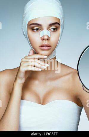 Belle femme après la chirurgie plastique, à la recherche dans le miroir. La photo d'une femme enveloppée dans des bandages médicaux. Beauté concept Banque D'Images