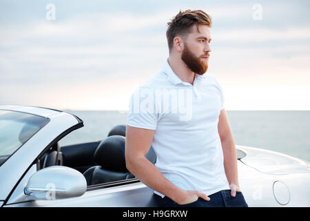 Beau barbu en chemise blanche leaning on car en étant debout sur la plage Banque D'Images