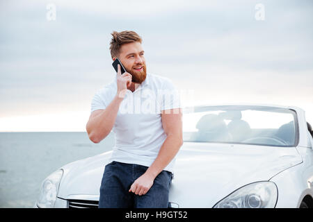 Beau jeune homme en conversation sur le téléphone mobile tout en se penchant sur sa voiture Banque D'Images