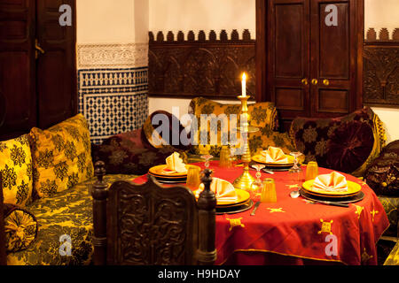 La lumière des bougies illumine une table dressée avec des serviettes roses dans un salon du Riad Arabesque à Fès, au Maroc, des portes en bois sculpté et des carreaux zellige bleus et blancs encadrent l'espace assis. Banque D'Images
