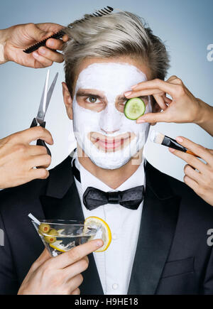 Bel homme avec masque facial hydratant entouré par les mains de service différents (styliste, coiffeur, coiffure). Photo de heureux homme élégant r Banque D'Images